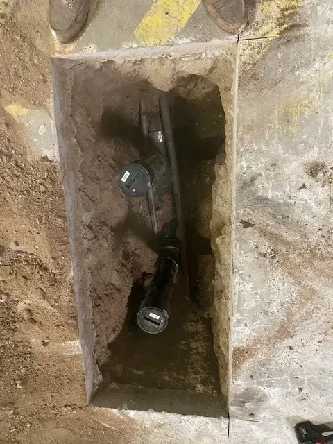 A rectangular trench cut into concrete reveals black pipes with black caps. The trench sits on a floor marked with yellow tape.
