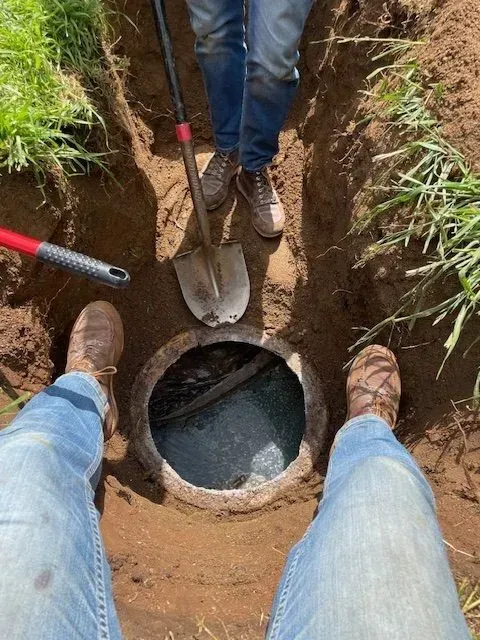 People digging around a manhole in a dirt area. One person is holding a shovel, and two pairs of legs are visible.