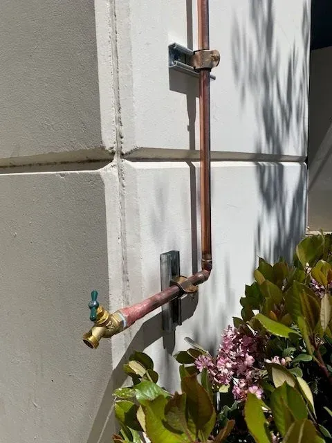 Copper water pipe with a spigot attached to a stucco wall, held in place by metal brackets.  Pink flowers and green foliage are in the lower right.