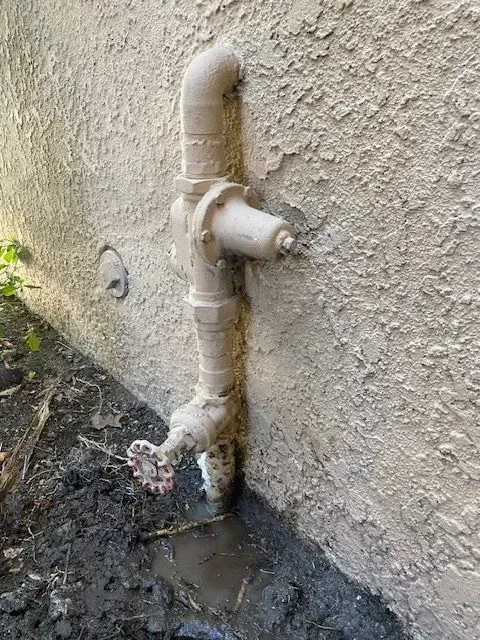 A weathered outdoor water pipe system on a stucco wall, with a valve and visible water leak at the base, sitting in muddy soil.
