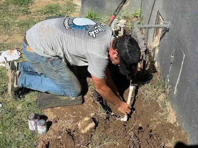 A man kneeling in dirt, wearing a gray shirt and jeans, connects white PVC pipe. He is working outside next to a dark wall.