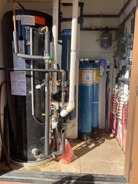 A utility room with a water heater, filtration system, and various pipes. The room has white and blue equipment, and a wooden floor.