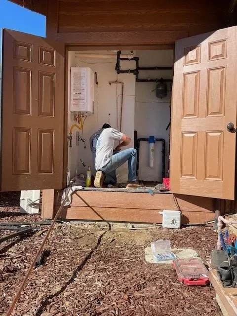A person installs plumbing in a small utility room with open doors. Brown doors and wood frame contrast with the white walls and equipment inside.