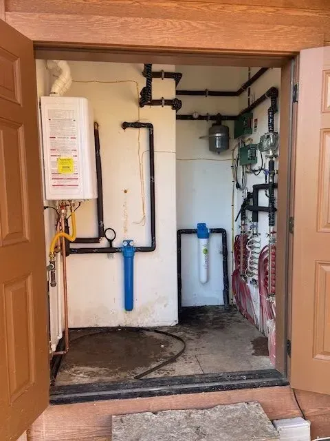 Interior of a utility room showing plumbing and heating equipment against white walls, accessible through open brown doors.