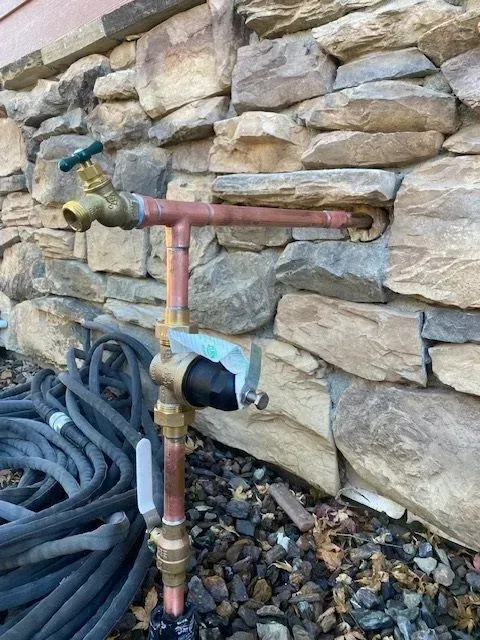Copper water pipe and outdoor faucet protruding from a stone wall; a coiled garden hose rests nearby.