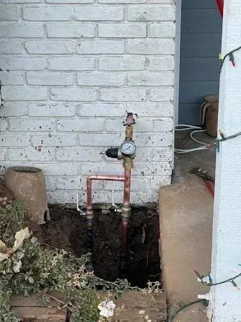 Copper pipes with a gauge and valve are installed outside near a white brick wall. A hole is dug around the pipes.