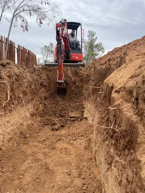 A small excavator digs a trench in red soil, with a person operating it. The work is being done near a wooden fence.