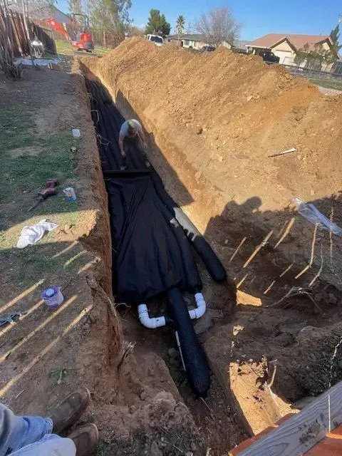 A narrow trench with black drainage fabric and white pipes laid in the dirt; person working nearby.