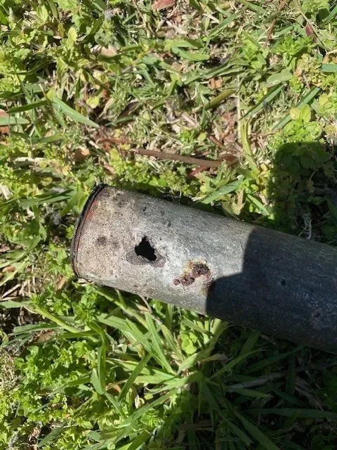 Close-up of a weathered, metallic pipe with a hole in its side, lying on green grass.