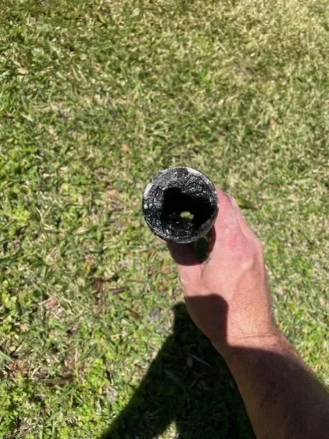A person's hand holds a black, open-ended pipe, likely plastic, with a hole inside; green grass in the background.