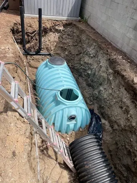 A light-blue septic tank sits in a dirt trench near a fence and house foundation. A ladder and black corrugated pipe are nearby.