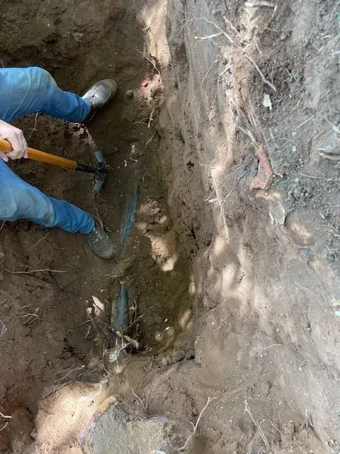Person digging in a trench, using a pickaxe, visible legs and arms, the dirt is dark, the setting is outdoors.