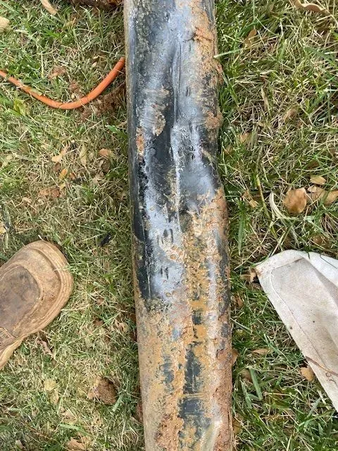 Black, dented pipe lying on green grass with dirt and a boot visible nearby.