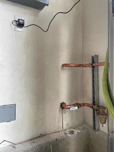 Copper pipes and a water valve on a wall. A black device with a cord is plugged into an outlet.