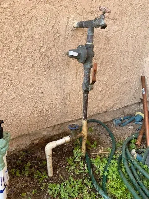 Exterior water pipes and spigot on a stucco wall, with green garden hose and weeds.