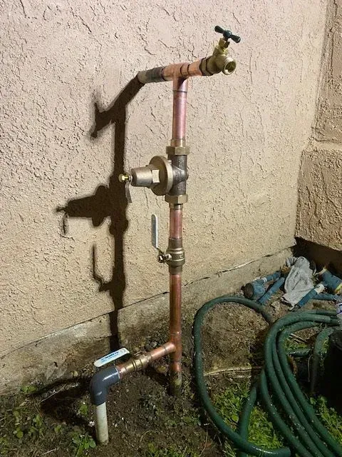 Copper pipes with faucets and a water pressure regulator, installed outside on a beige stucco wall, casting a shadow on the ground.