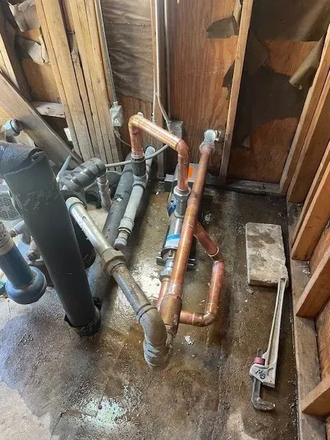 Plumbing pipes in a cluttered space. Copper and metal pipes are present, with a wrench leaning against a wall. The floor is wet.