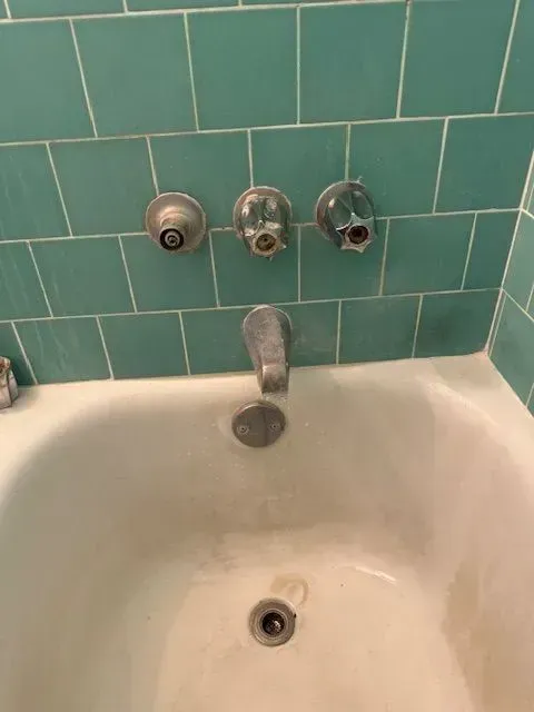 Bathtub with teal tiled wall and three faucet handles, one with a faucet and drain.