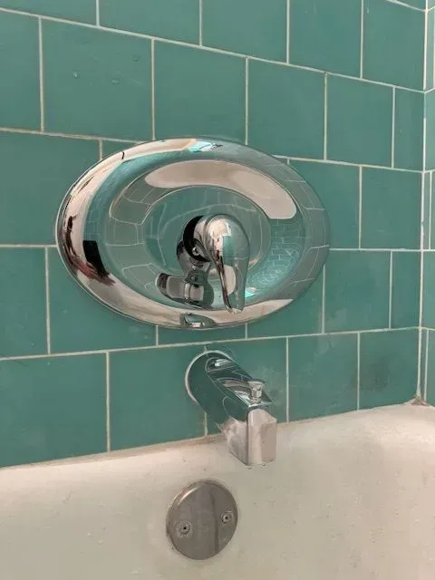 Chrome faucet and shower handle on a teal tiled wall above a white bathtub.
