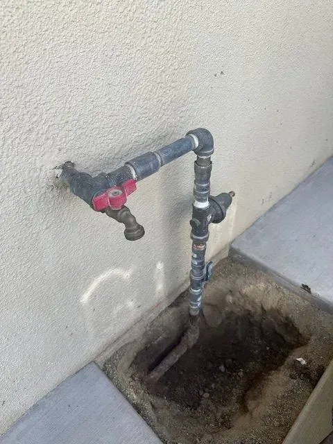 A water pipe assembly with a faucet on an exterior wall. The pipes are black and gray, with a red-handled valve. The assembly sits above a recessed area.