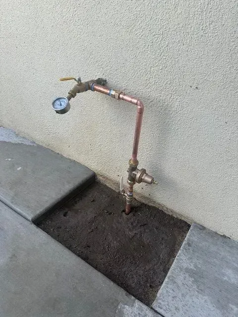 Copper plumbing fixture with a pressure gauge and valve, mounted near a concrete sidewalk and beige wall. It is installed in a patch of exposed soil.