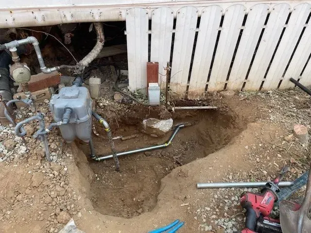 A trench dug near a house foundation, revealing gas and water pipes. A worker's tools, including a drill, lie nearby.