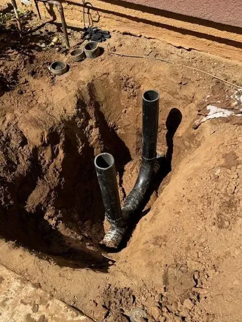 Black plumbing pipes emerge from a dug-out pit in the dirt. The pipes have a Y-shaped configuration.