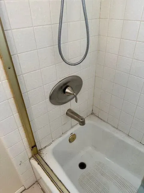 A bathtub and shower with white tiles. The water controls are silver, and a handheld showerhead hangs above.