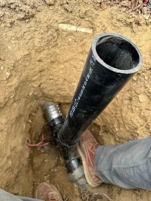 A person in jeans and work boots standing in a dirt pit, with a black plumbing pipe and fittings exposed.