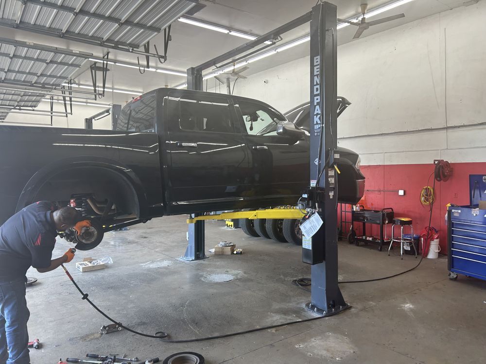 Black pickup truck on a lift in an auto shop, with a mechanic working underneath Graphic  Image | All Tune Total Car Care