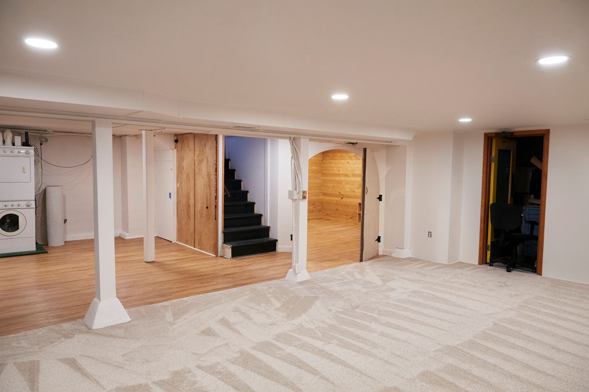 Finished basement with white walls, wood floors, carpet, support beams, doors, and stairs.