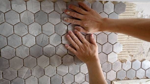Hands installing hexagon-shaped gray tiles on a wall.