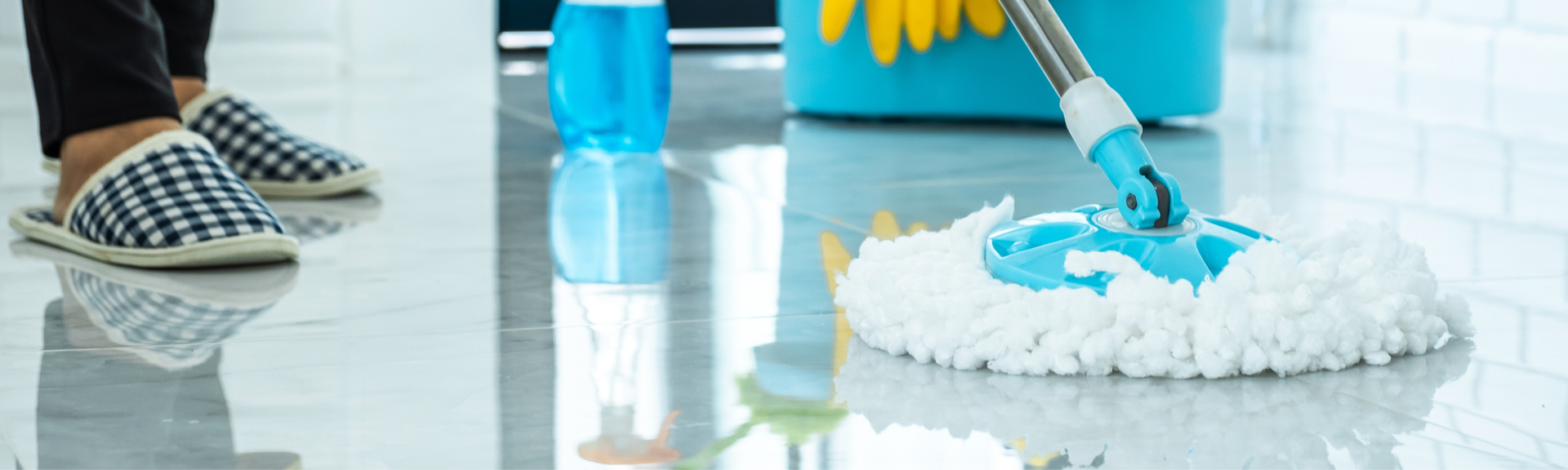 A person in plaid slippers mopping a glossy tile floor with a blue mop, with a spray bottle and bucket nearby.