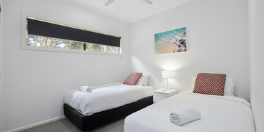 A bedroom with two twin beds featuring white bedding, dark headboards, patterned pillows, and a window with a dark shade.