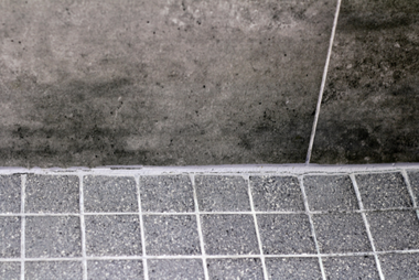 A close-up of a shower corner showing a gray, speckled tiled floor meeting a textured, dark gray wall with visible mold.