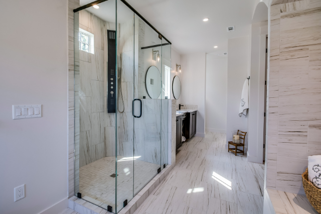 A modern bathroom with a glass-enclosed shower, light marble tile walls and floors, a vanity, and natural lighting.