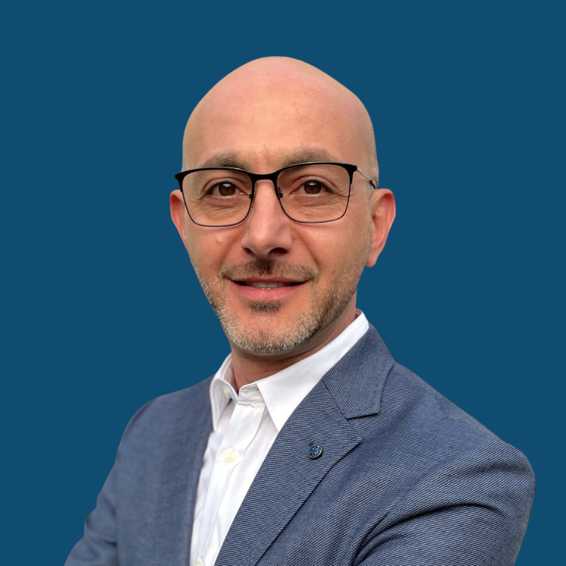 Man with glasses, wearing a blue blazer and white shirt, smiles against a blue background.