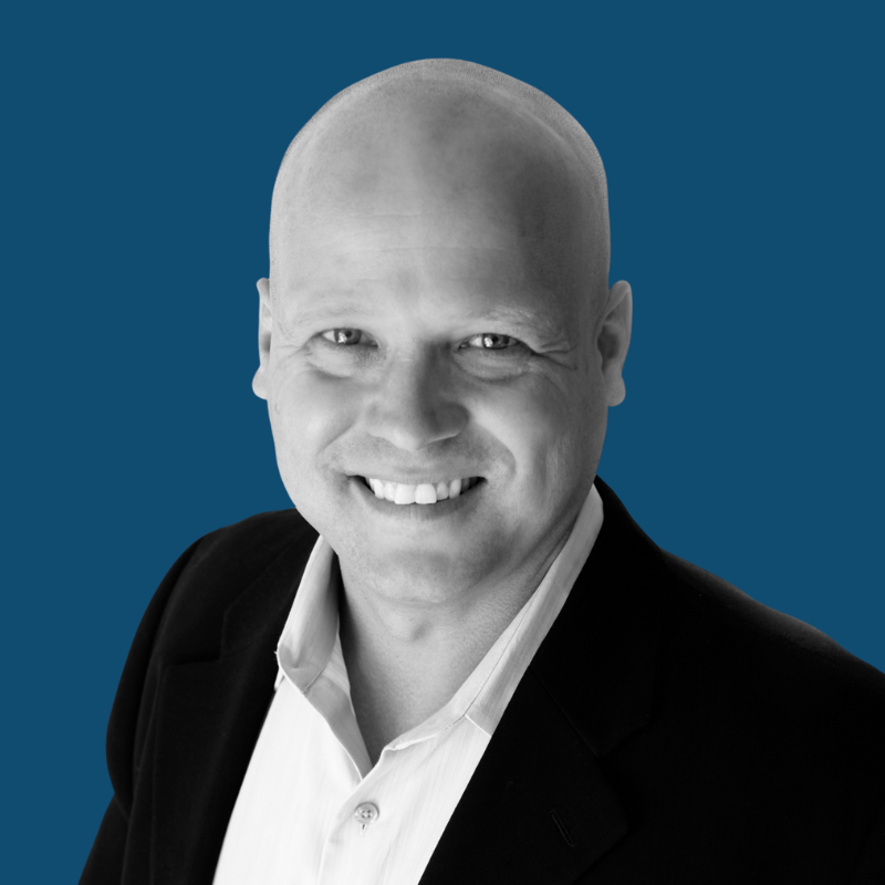 Bald man smiles, wearing a black blazer and white shirt against a blue background.