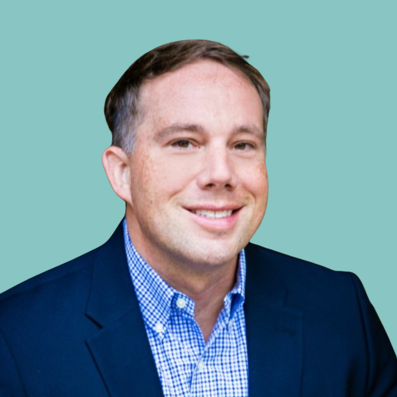 Man in a navy blazer and blue checkered shirt smiles against a light blue background.