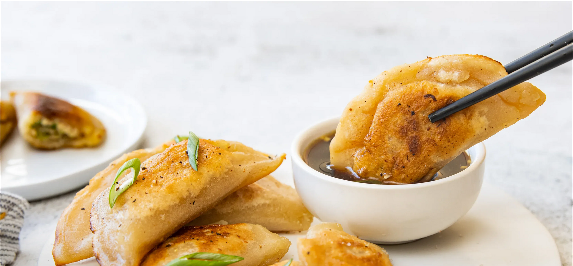 Gluten Free Dumplings being dipped in sauce, held by chopsticks. Several dumplings on a plate, green garnish.