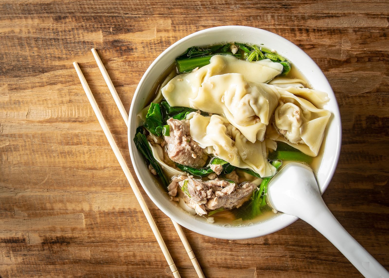 A bowl of pork wonton soup with chopsticks and a spoon on a wooden table.