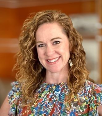 A woman with curly hair is smiling and wearing a colorful shirt.