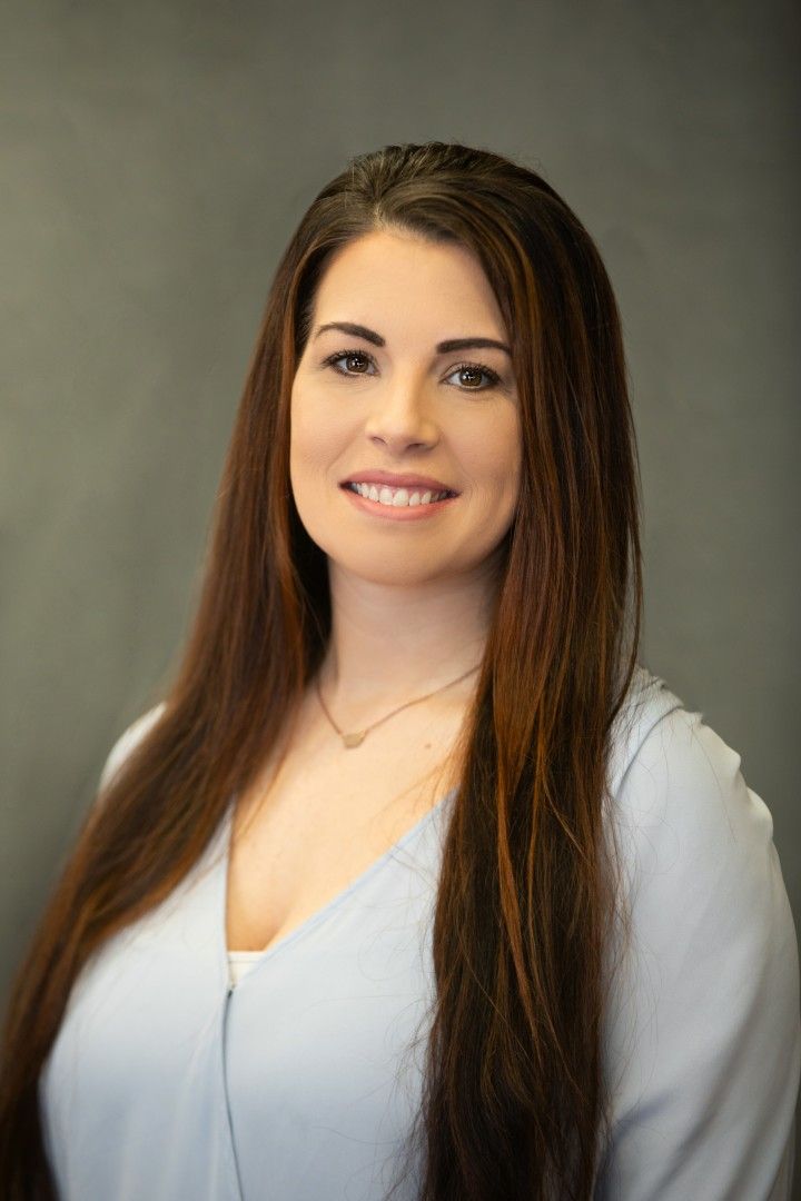 Portrait of a smiling woman with long brown hair in a light blue top against a gray background