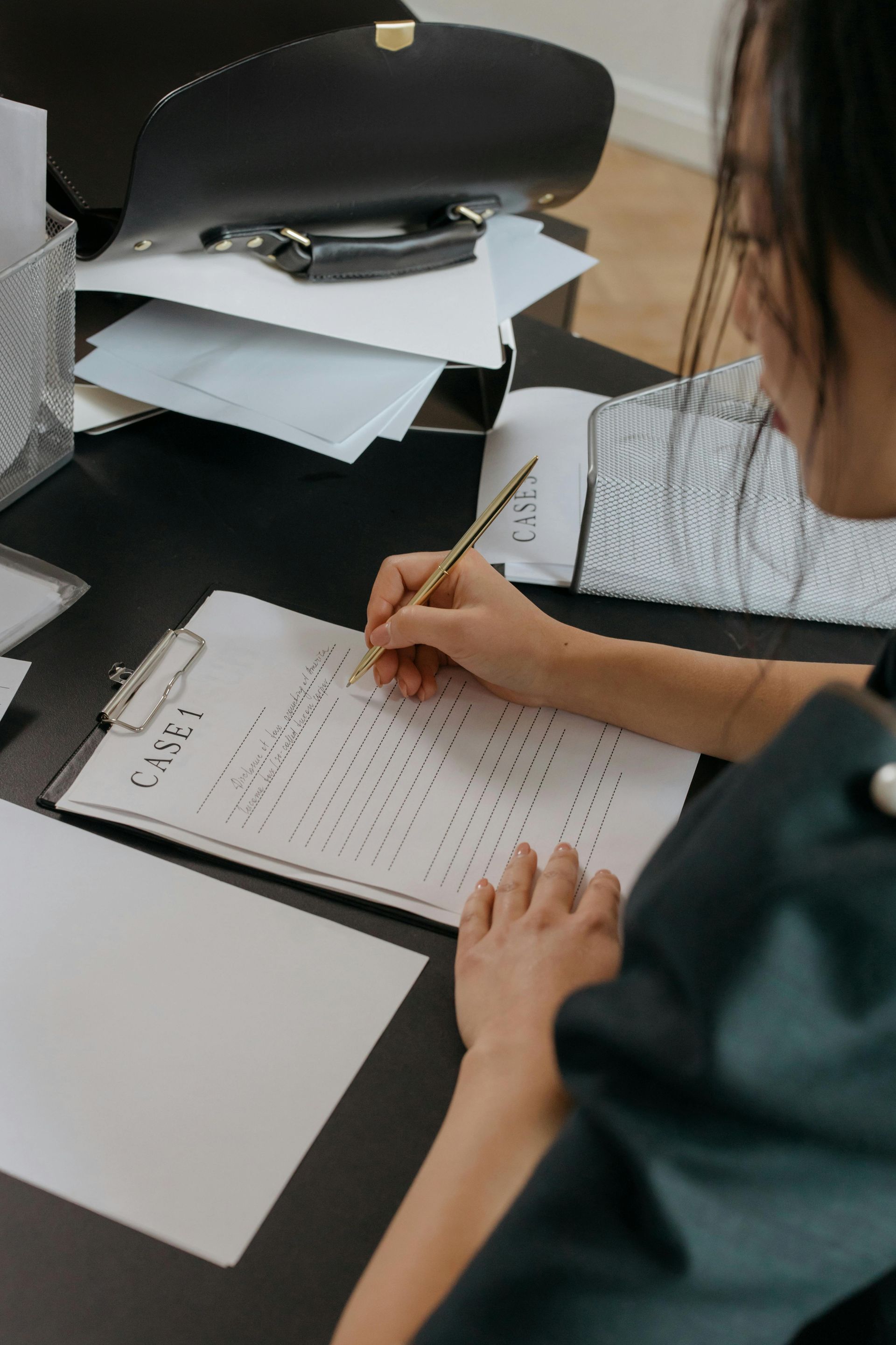 Person writing on a clipboard at a desk; papers and a purse are visible in the background.