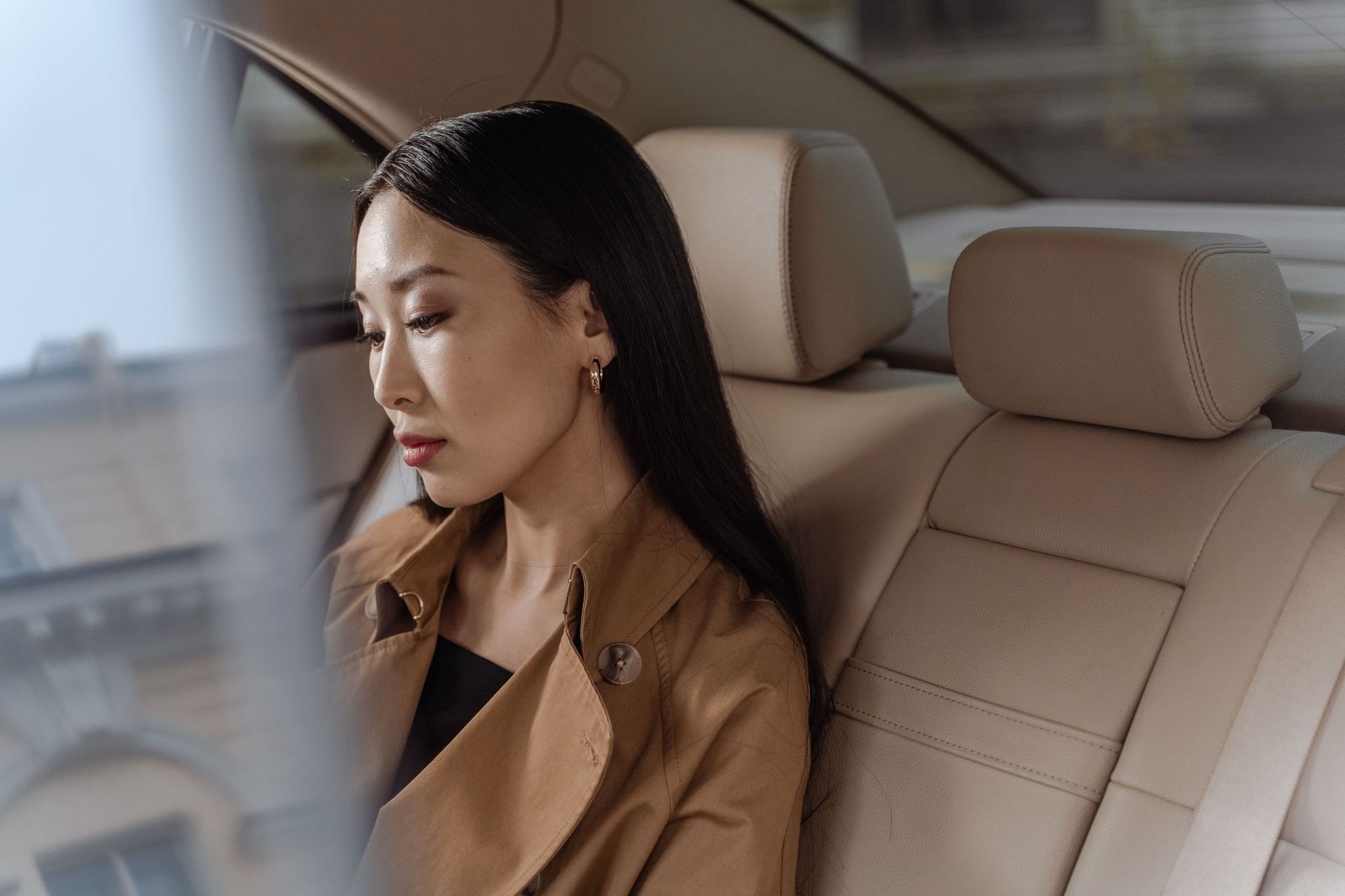 Woman in a tan coat sitting in the back seat of a car, looking out the window.