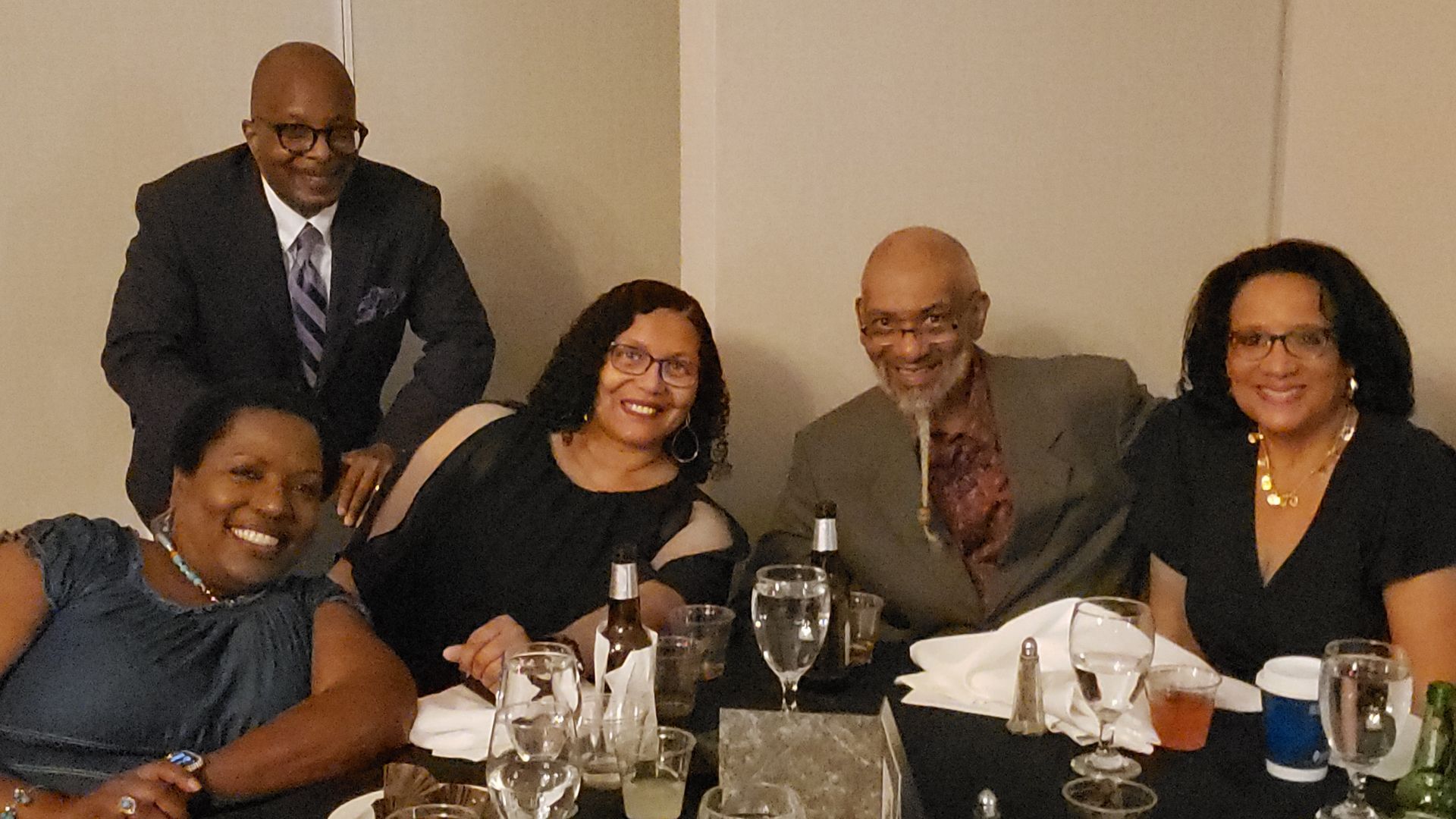 Five people seated at a table, smiling. A man stands behind them. Formal dinner setting.