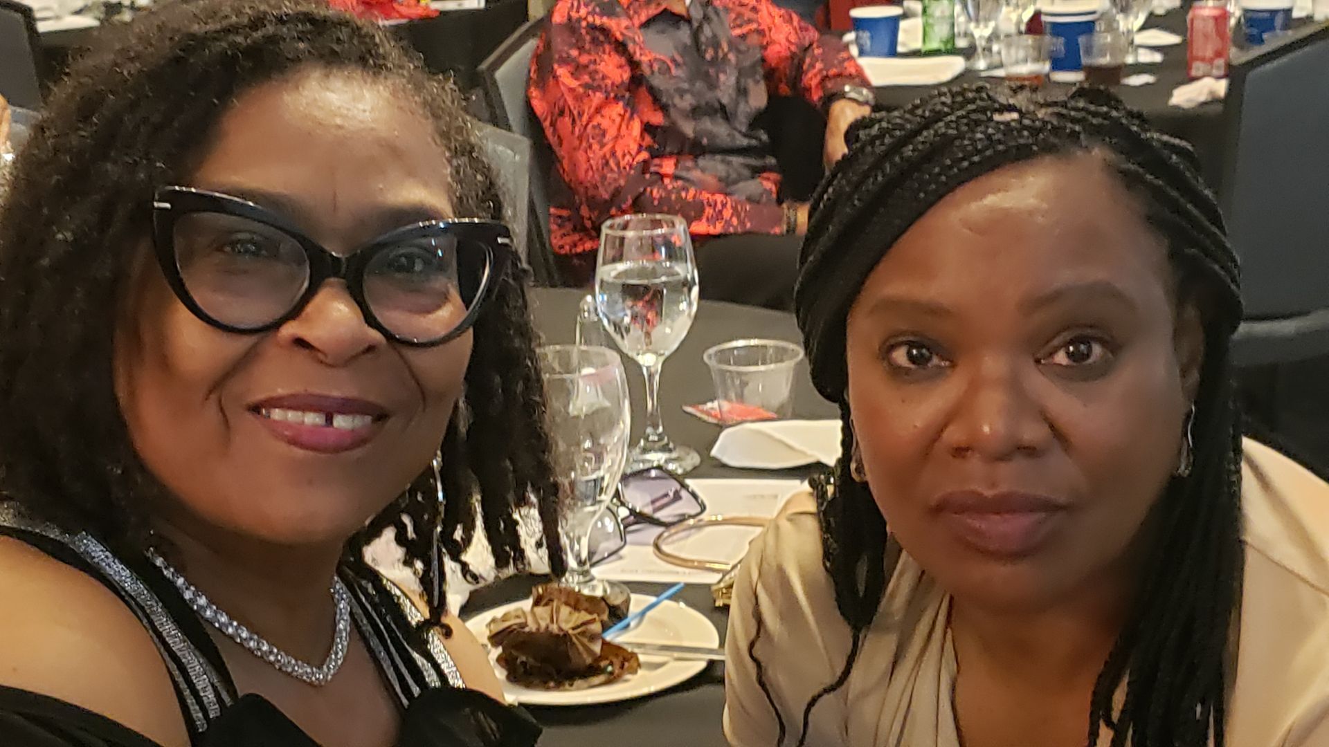 Two women smiling at a table, drinks, food; one in glasses, the other with braids, at a formal event.