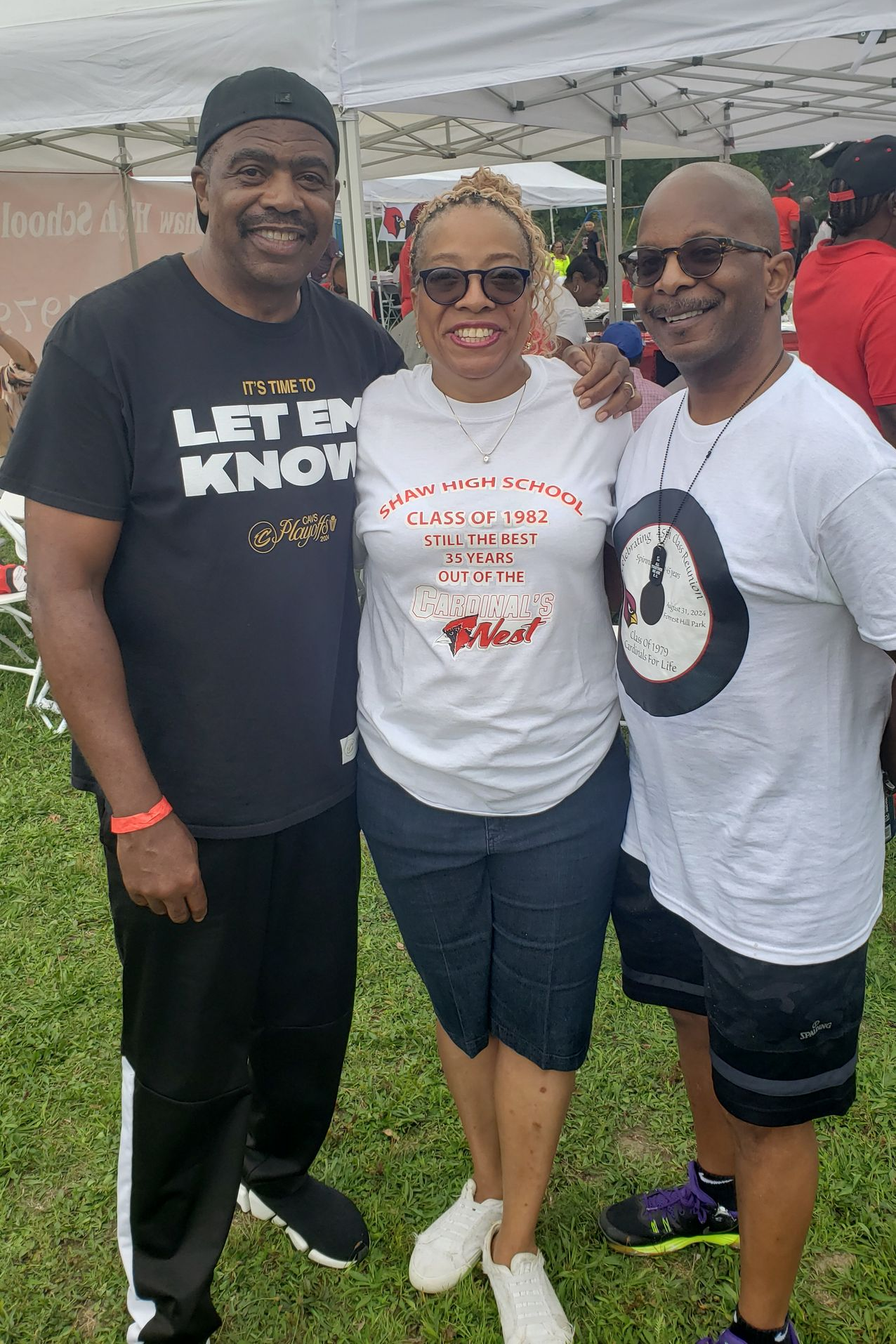 Three people smiling for a photo outdoors. Woman in center, surrounded by men. All are wearing casual clothes and sunglasses.