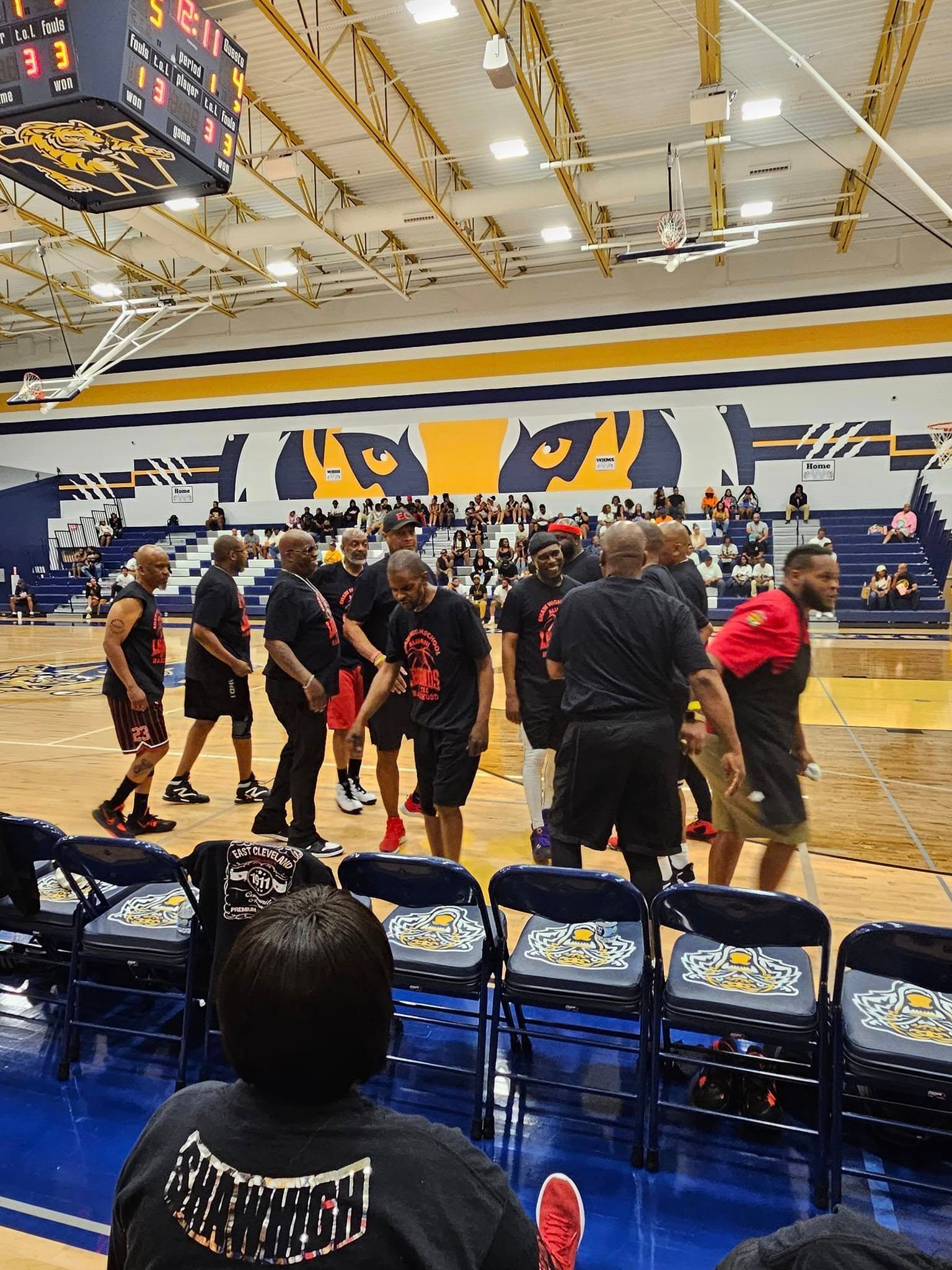 A group of men in black shirts stand on a basketball court, possibly a team. Spectators are visible in the background.