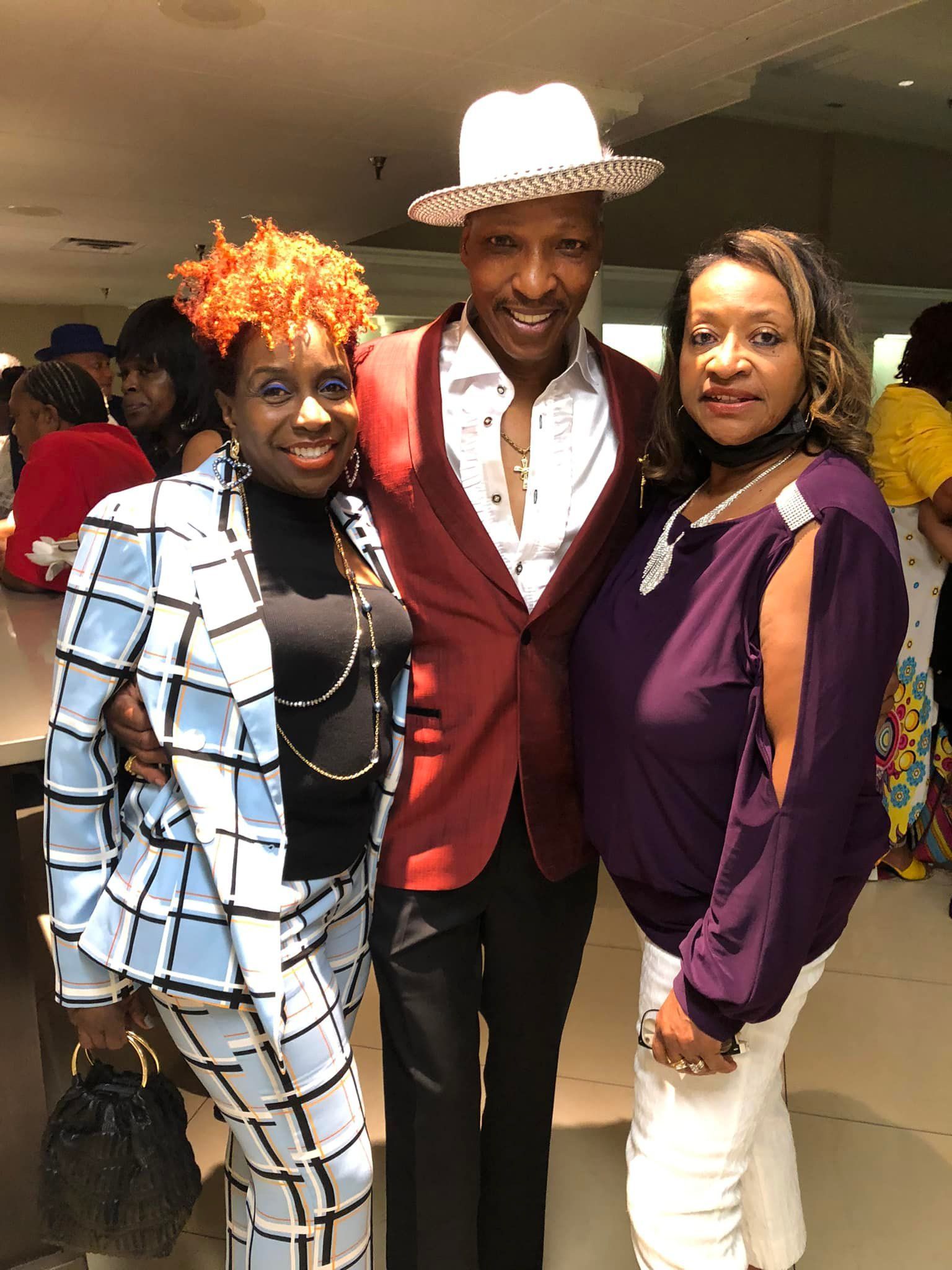 Three people posing: man in red jacket and hat, woman in blue suit, woman in purple top and white pants.